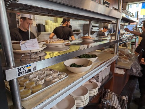 A busy kitchen with shelves displaying dishes, sauces, and staff preparing food in the background.
