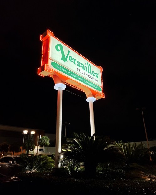 Neon sign for Versailles Cuban Cuisine, illuminated at night, surrounded by palm trees.