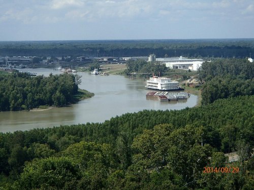 A scenic view of a river with boats, surrounded by lush greenery and industrial buildings in the background.