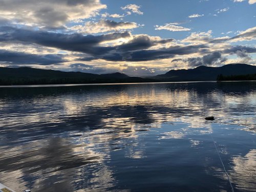 A serene lake at sunset, reflecting clouds and mountains in calm waters under a colorful sky.