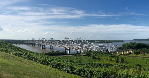 A panoramic view of a white steel bridge spanning a river, surrounded by green hills and a clear blue sky.