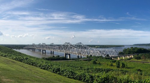A panoramic view of a white bridge spanning a river, surrounded by green hills and a clear blue sky.