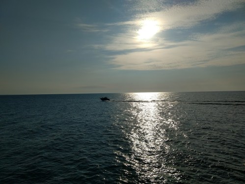 A boat glides across a shimmering ocean under a bright sun and partly cloudy sky.