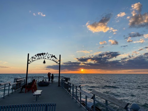 A couple embraces on a pier at sunset, with colorful clouds and the sun setting over the ocean in the background.