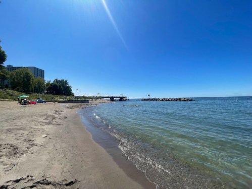 A sunny beach scene with clear blue water, sandy shore, and distant pier under a bright blue sky.