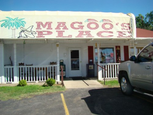 A small restaurant with a sign reading "Magoo's Place," featuring a porch and tropical decorations.