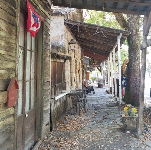 A rustic stone pathway lined with wooden buildings, chairs, and a small American flag, surrounded by trees and fallen leaves.