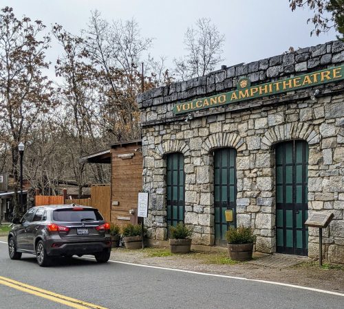A car drives past the stone entrance of Volcano Amphitheatre, surrounded by trees and a cloudy sky.
