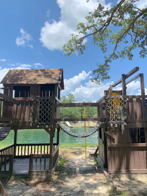 A wooden playground structure by a lake, featuring a tower, bridge, and swings under a blue sky with fluffy clouds.