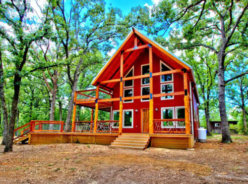 A modern red cabin with a large deck, surrounded by trees and blue skies.