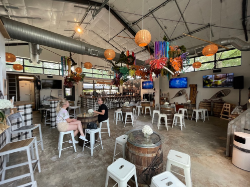 A spacious, modern bar with wooden barrels, high tables, and colorful decor, featuring two people seated at a table.