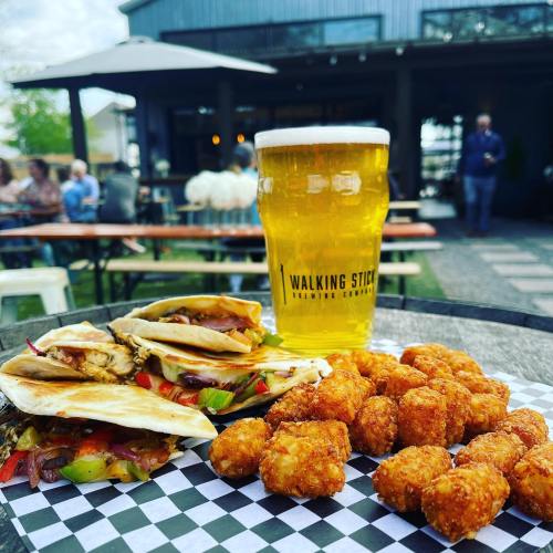 A plate of tacos and crispy tater tots beside a tall glass of beer, set outdoors at a lively restaurant.