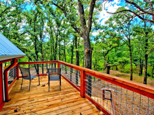 A wooden deck with two chairs overlooks a lush, green forest under a bright blue sky.