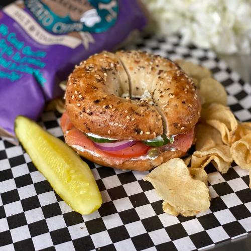 A bagel sandwich with smoked salmon, tomato, and onion, served with potato chips and a pickle on a checkered plate.