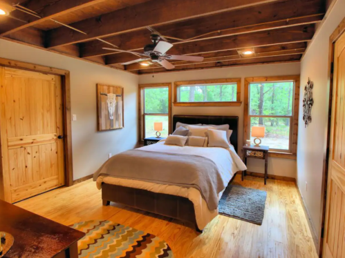 Cozy bedroom with wooden beams, a queen bed, two lamps, and large windows overlooking greenery.