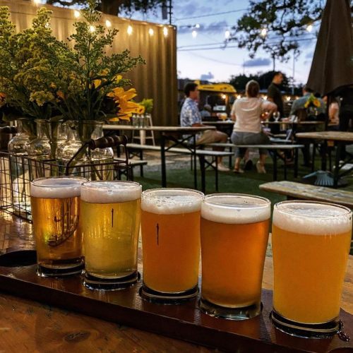 A flight of five craft beers on a wooden table, with a lively outdoor setting and people in the background.