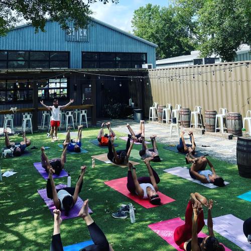 A group of people practicing yoga outdoors on mats, with a teacher leading in front of a rustic building.