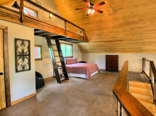 Cozy loft bedroom with wooden beams, a double bed, and a ladder leading to a lower level. Warm, inviting atmosphere.