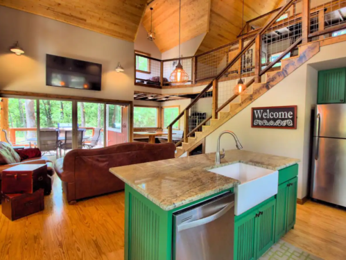 Cozy cabin interior with wooden beams, a kitchen island, and a welcoming sign, featuring large windows and a staircase.