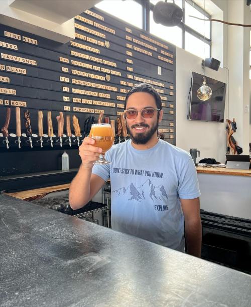 A smiling man in a light blue shirt holds a beer glass at a bar with a wooden tap wall in the background.