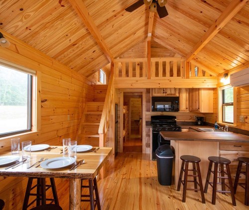 Cozy wooden cabin interior with a dining table, kitchen, and lofted sleeping area, featuring warm wood tones and natural light.