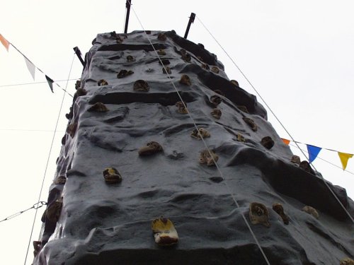 A tall rock climbing wall with various holds, surrounded by colorful flags and a cloudy sky.