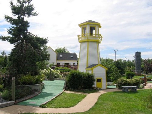 A yellow and white lighthouse stands in a mini-golf course, surrounded by greenery and a blue sky.