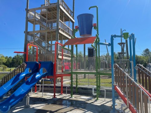 Colorful playground structure with slides, water features, and a large bucket, set against a clear blue sky.