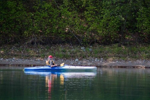 A person in a kayak paddles on calm water near a lush green shoreline.
