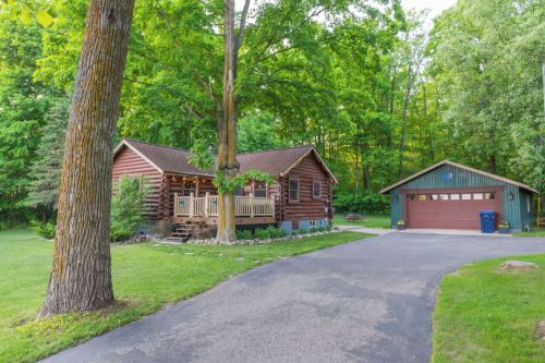 A charming log cabin surrounded by trees, with a driveway leading to a garage and a front porch.