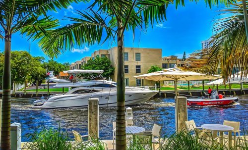A scenic view of boats on a canal, surrounded by palm trees and outdoor seating. Bright blue sky and buildings in the background.