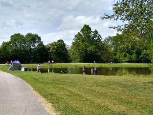 A serene park scene with a pond, people fishing, and lush greenery under a partly cloudy sky.