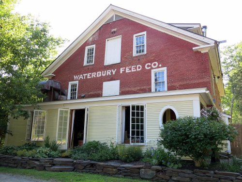 Historic building with red brick and yellow siding, featuring "Waterbury Feed Co." painted on the front. Lush greenery surrounds it.