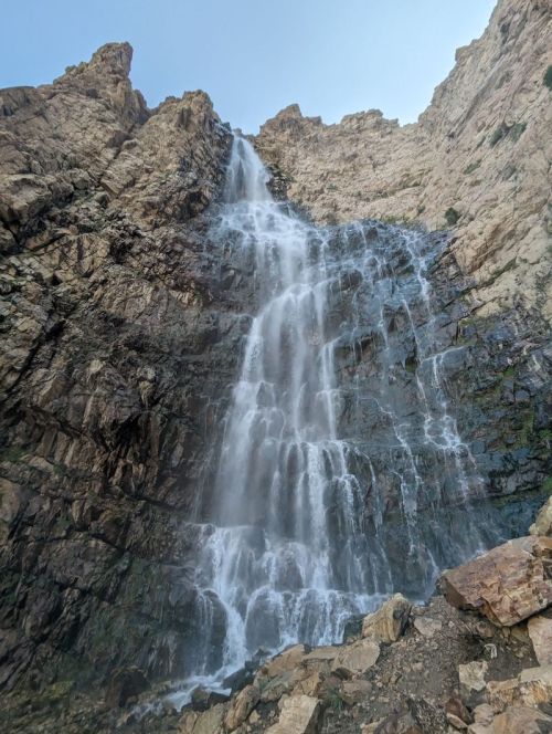 A tall waterfall cascading down a rocky cliff, surrounded by rugged terrain and clear blue sky above.