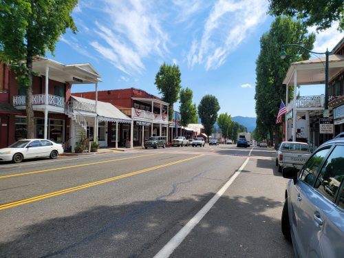 A quiet street in a small town, lined with shops and trees, under a blue sky with scattered clouds.