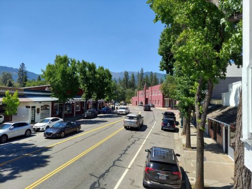 A sunny street scene with trees, parked cars, and shops lining the road, set against a backdrop of mountains.