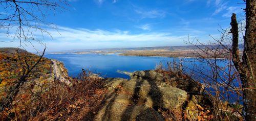 A scenic view of a river surrounded by autumn foliage and blue skies, with rocky terrain in the foreground.