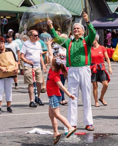 A performer in a green shirt creates a large bubble as a girl in a red shirt reaches for it in a lively outdoor setting.