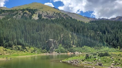 A serene mountain landscape featuring a calm lake surrounded by lush green trees and rocky hills under a partly cloudy sky.