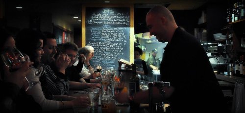 A bartender serves drinks to patrons at a lively bar, with a chalkboard menu in the background.