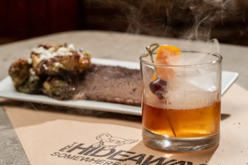 A cocktail with smoke, garnished with a cherry and orange, beside a plate of grilled meat and roasted vegetables.
