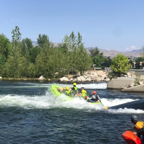 A group of people in a bright green raft navigates whitewater rapids, surrounded by trees and rocky banks.