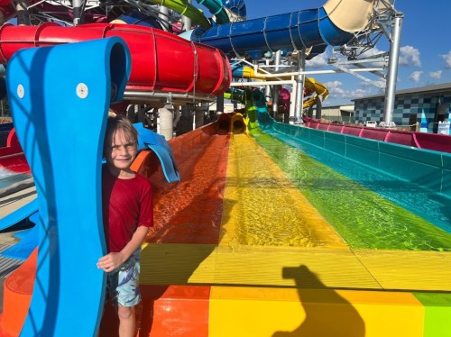 A child stands beside colorful water slides at a water park, smiling in the bright sunlight.