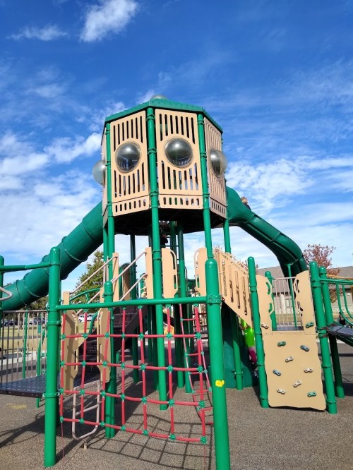 A colorful playground structure with slides, climbing walls, and a tower under a blue sky with scattered clouds.