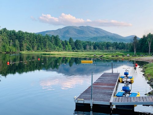 A serene lake scene with a wooden dock, pedal boats, and mountains in the background under a clear blue sky.