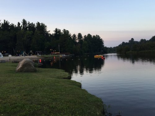 A calm lake at dusk, surrounded by trees, with a small boat and a rocky shore in the foreground.