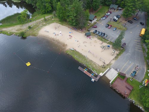 Aerial view of a sandy beach by a lake, with people swimming, a parking lot, and a dock with boats.