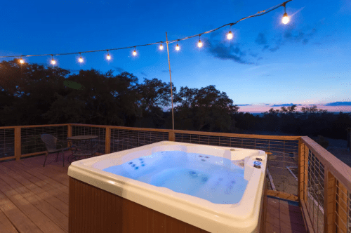 A hot tub on a wooden deck, illuminated by string lights, with a twilight sky in the background.