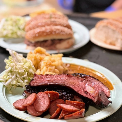 A plate of smoked brisket and sausage with sides of coleslaw and macaroni, with sandwiches in the background.