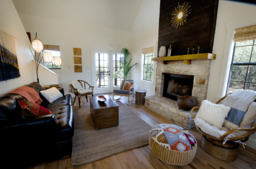 Cozy living room with a stone fireplace, wooden furniture, and natural light from large windows. Warm, inviting decor.